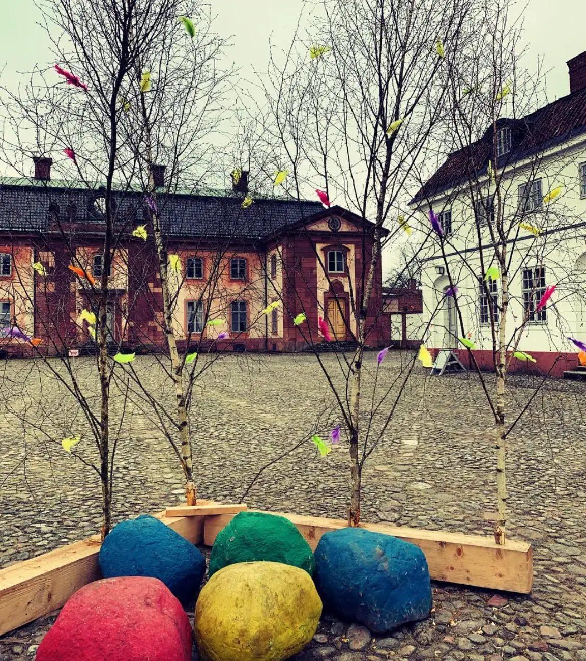 Påskris av nakna björkkvistar med färgglada fjädrar och stora målade ägg i en låg träram på kullerstensborggården vid Övedskloster