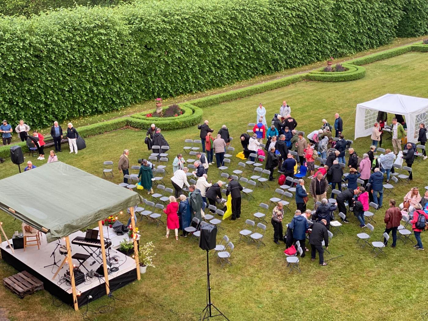 Utomhusevent på gräsmattan i parken med scen, rader av stolar och ett vitt tält framför en hög klippt häck