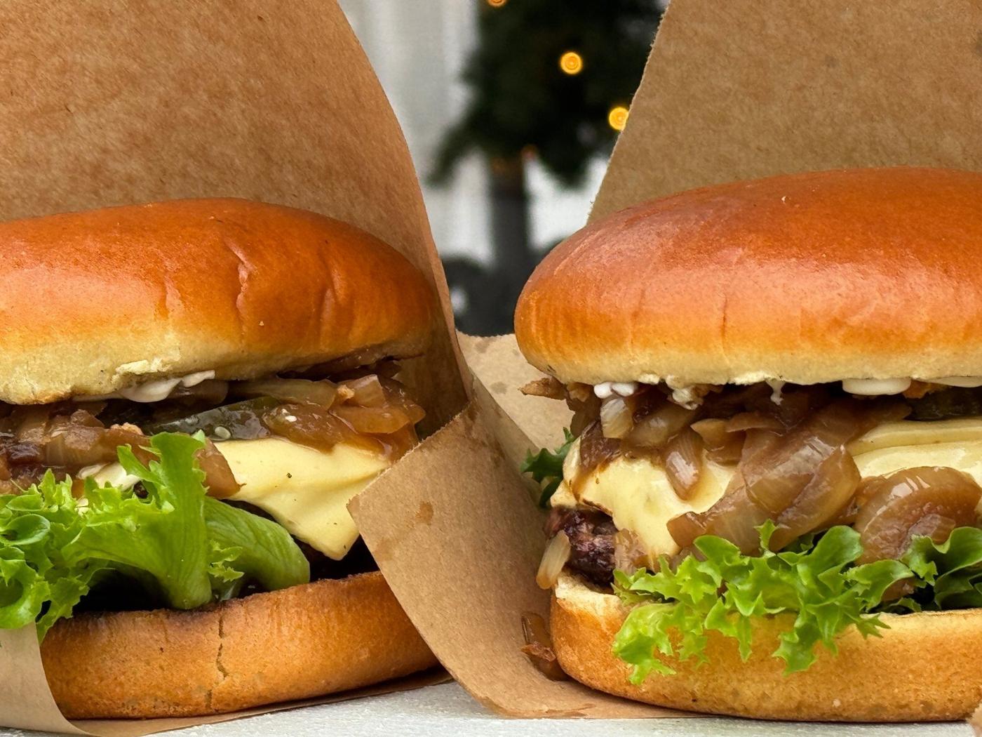 Två hamburgare i pappersfickor med ost, karamelliserad lök och sallad, med gran och ljusslinga i bakgrunden