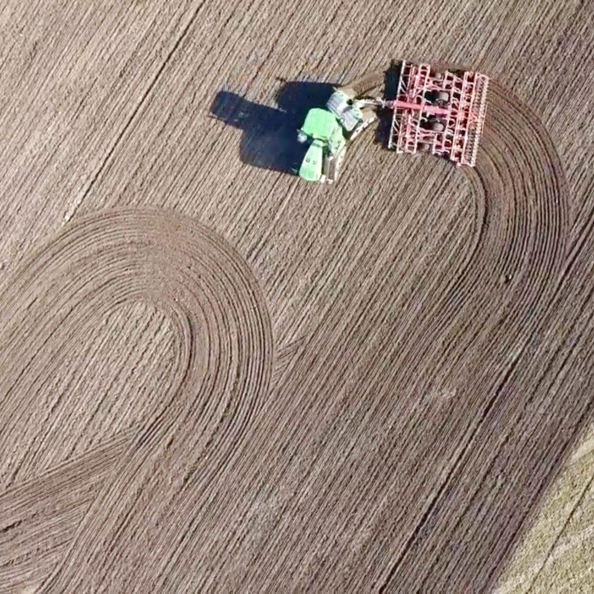 Åker sedd ovanifrån där en grön traktor drar en röd harv som bearbetar jorden i svepande spår