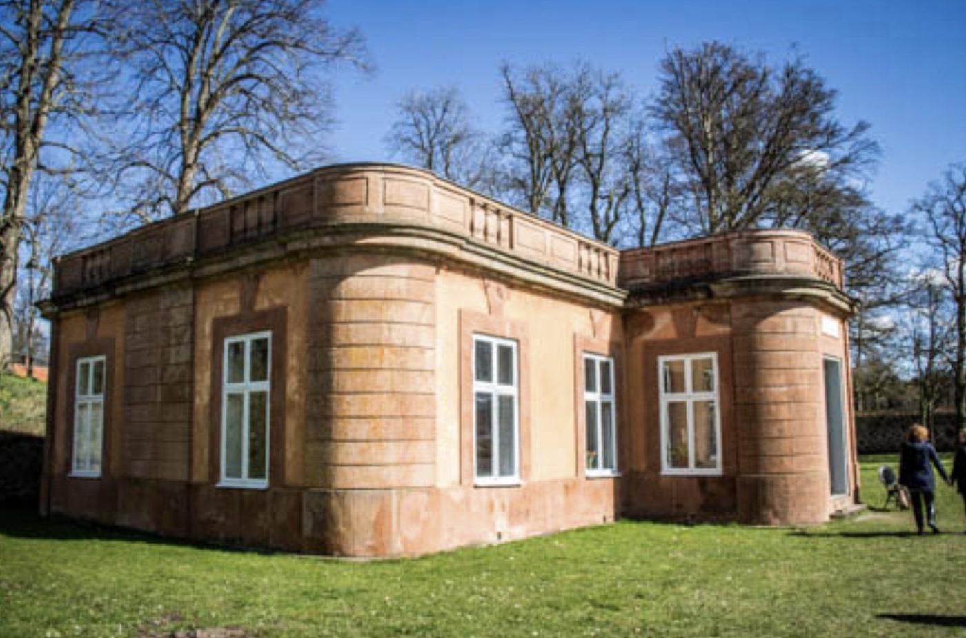 Volièrens putsade paviljong i slottsparken, med rundade hörn, balustrad på taket och höga vitmounterade fönster