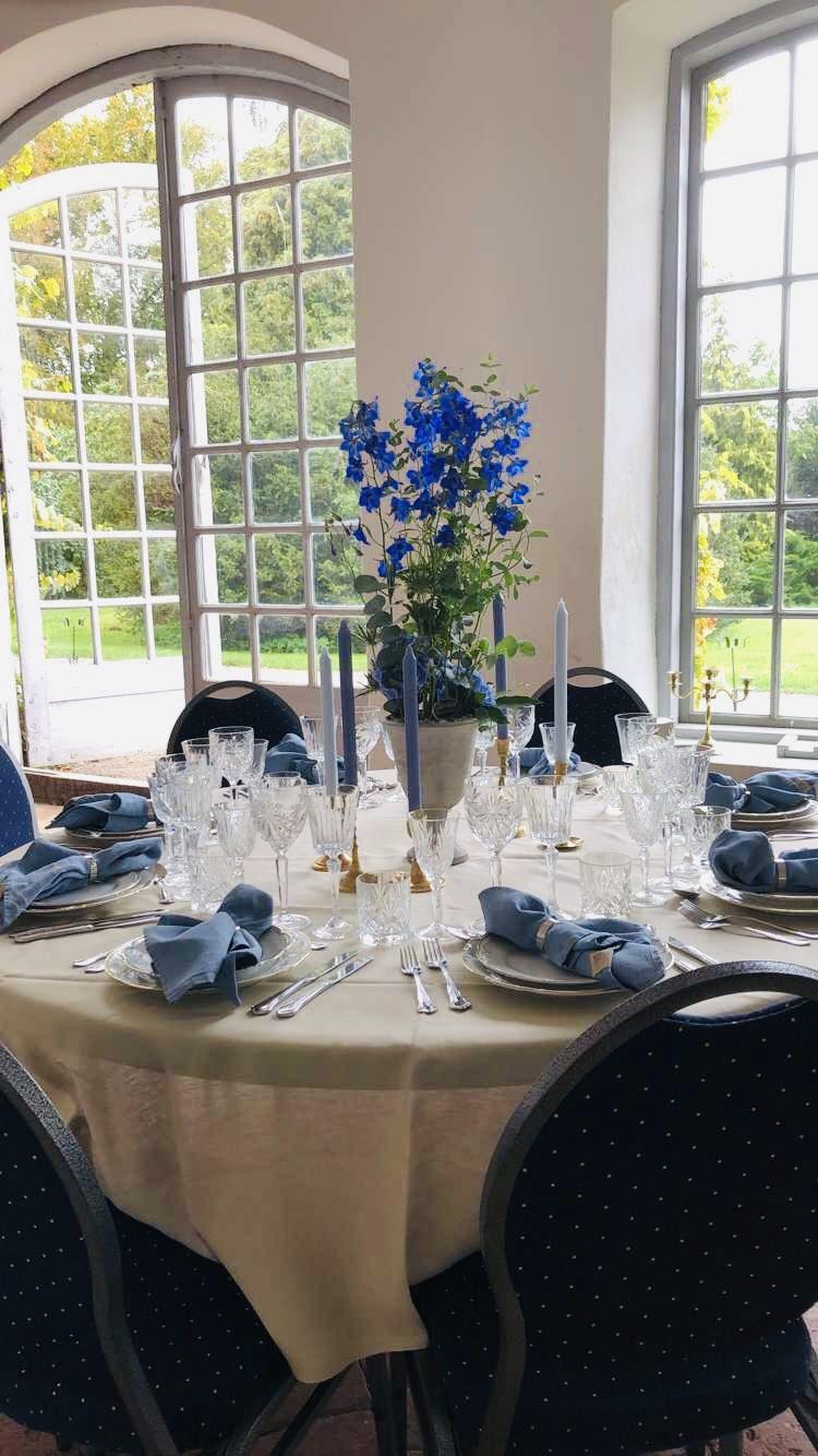 Dukat festbord i Orangeriet med vita dukar, blå servetter, blå ljus och en hög vas med blå blommor framför de höga fönstren