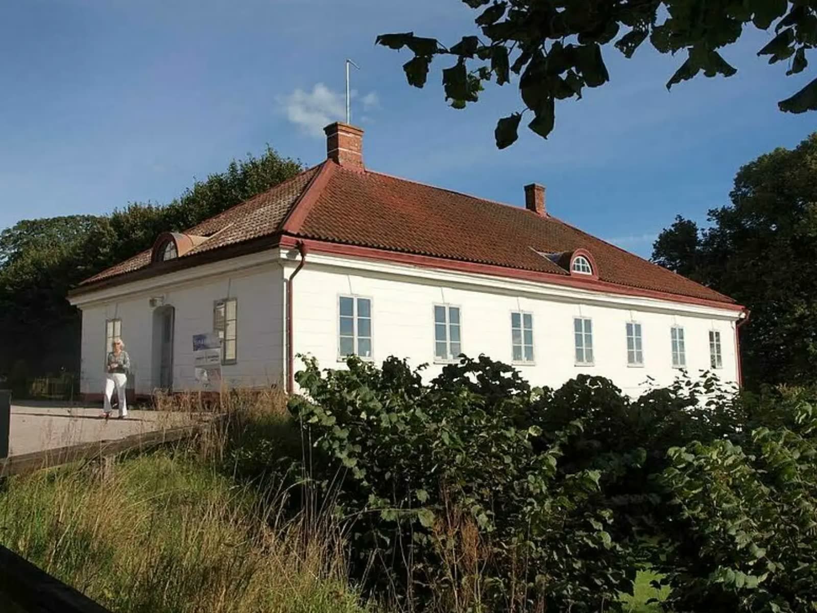 Skolhuset på Övedskloster, en vit träpanelad byggnad med rött tegeltak, omgiven av uppvuxna träd och buskar i sol