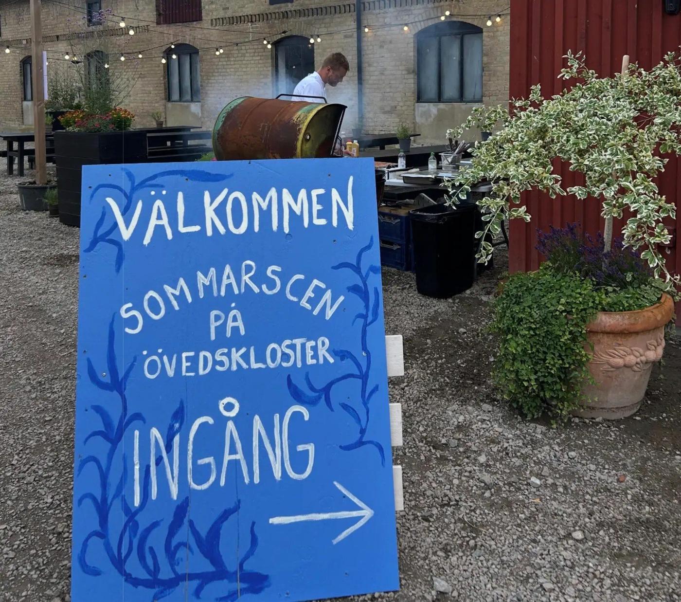 Handmålad blå välkomstschild för Sommarscen på Övedskloster med grillstation och personal i bakgrunden